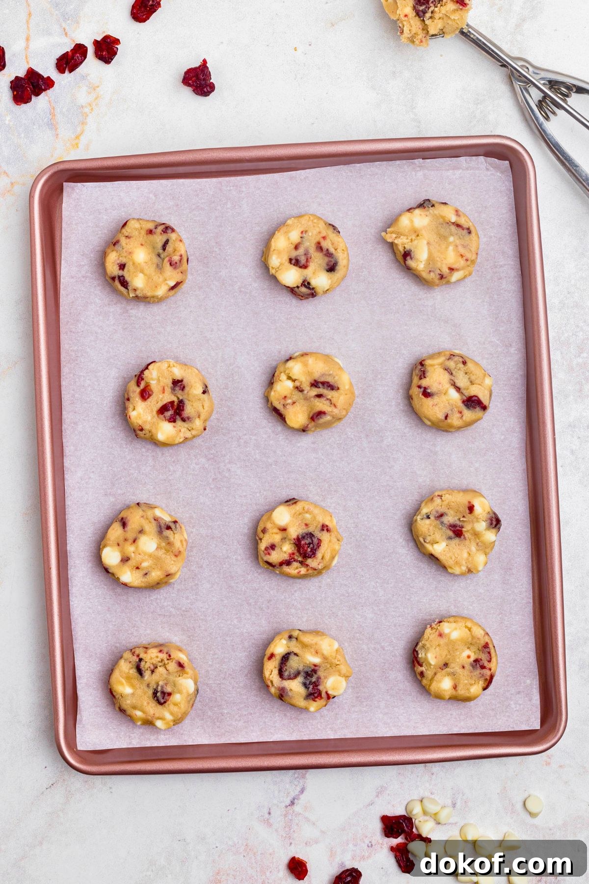 Balls of cookie dough dropped onto a baking sheet with cranberries and chips scattered around the table. 