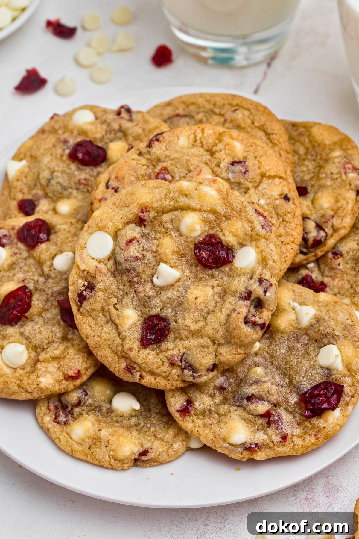 White chocolate chip and cranberry cookies stacked on a white plate with glasses of milk on the table.