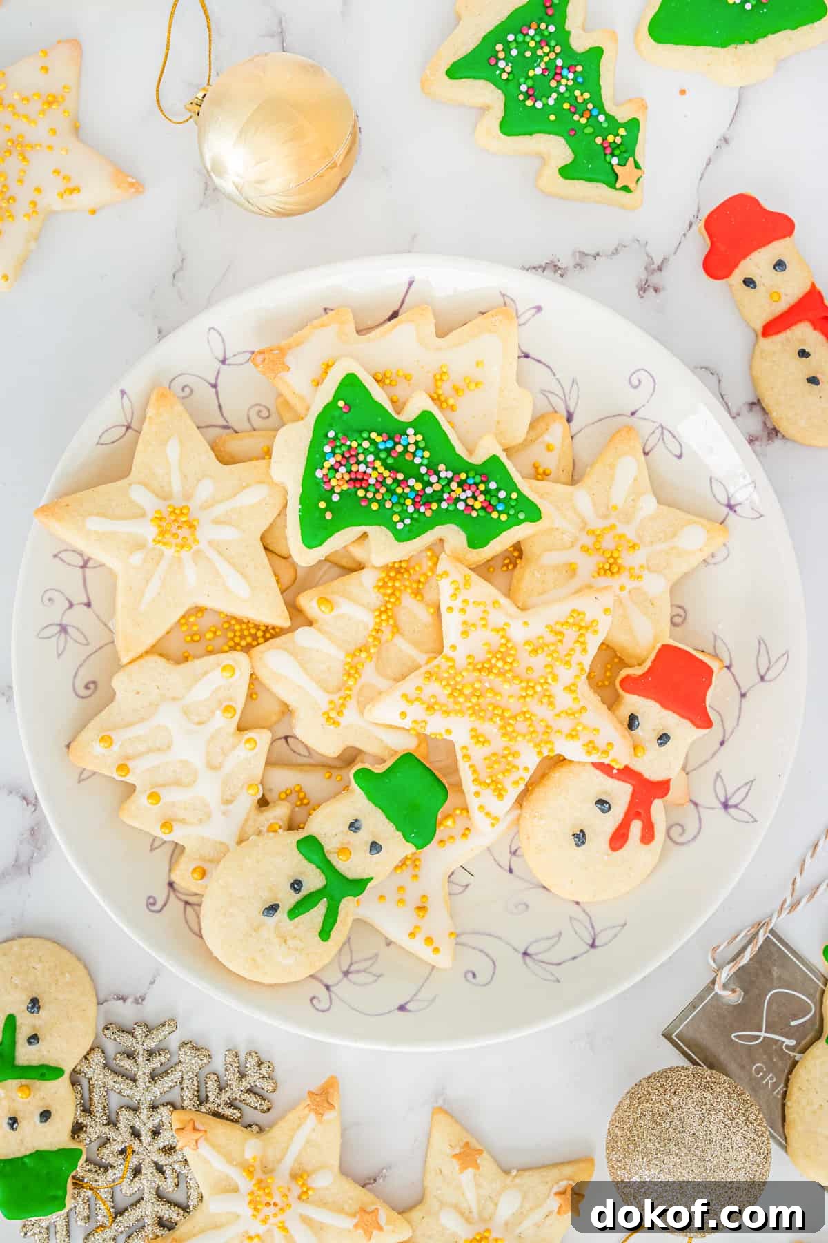 Decorated Christmas sugar cookies with icing and sprinkles on serving plate with additional cookies and Christmas ball ornaments scattered around table