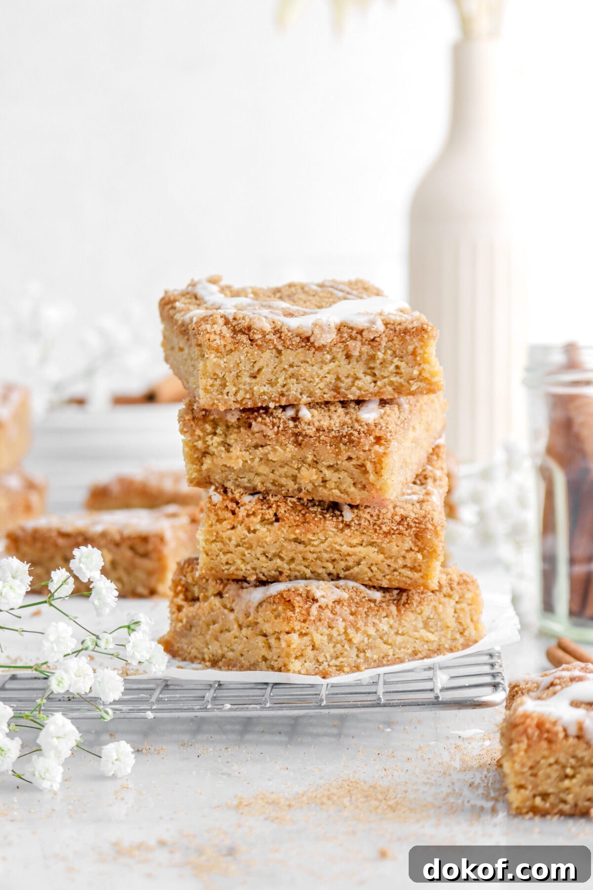 four stacked cinnamon roll blondies in wire cooling rack with white flowers beside, more blondies around, and a vase behind.