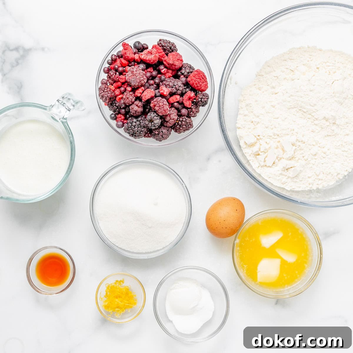 A top-down view of bowls containing flour, mixed berries, sugar, milk, melted butter, an egg, vanilla extract, lemon zest, and baking soda on a white surface.