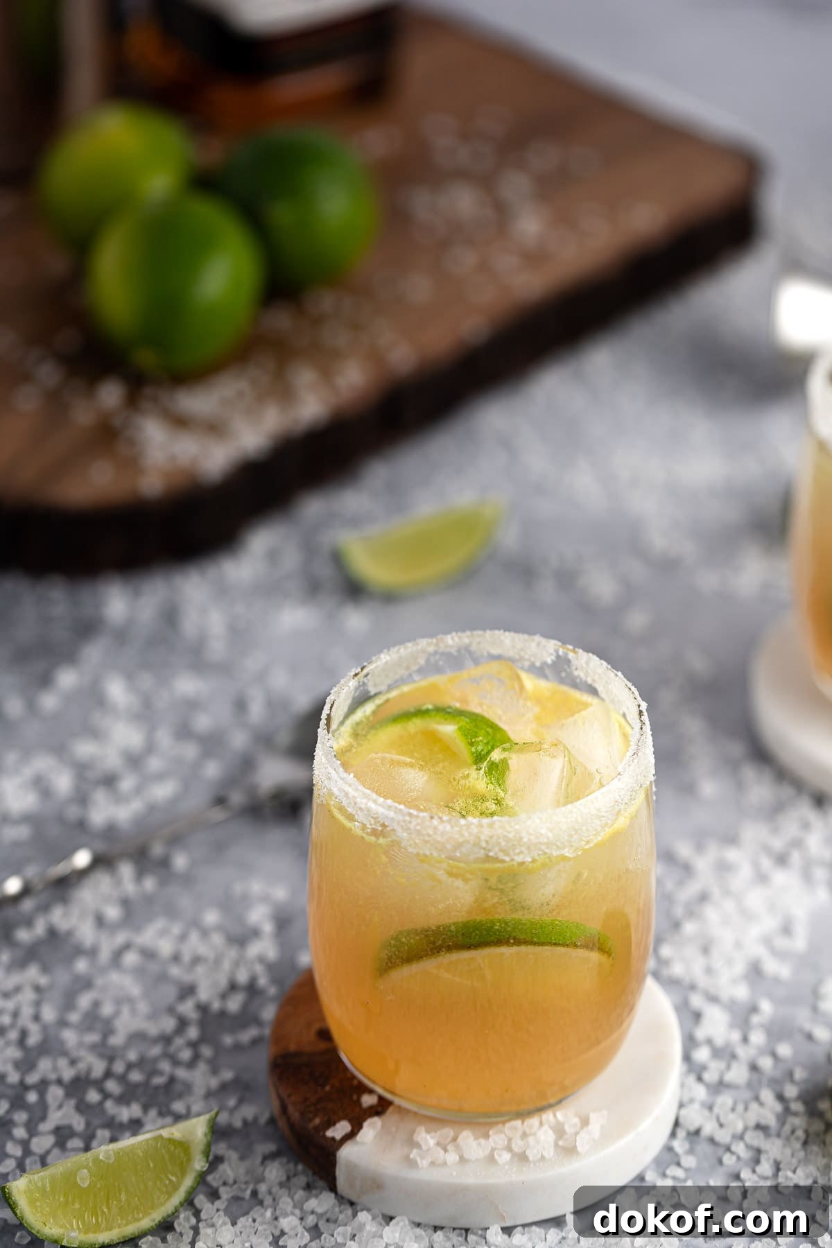 Bourbon margarita on a white and wooden coaster, with course salt scattered on the table, and a wooden serving board in the background.