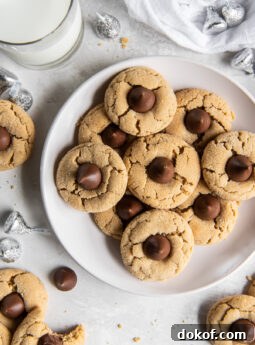 plate of peanut butter blossoms with chocolate kisses next to unwrapped kisses and glass of milk.