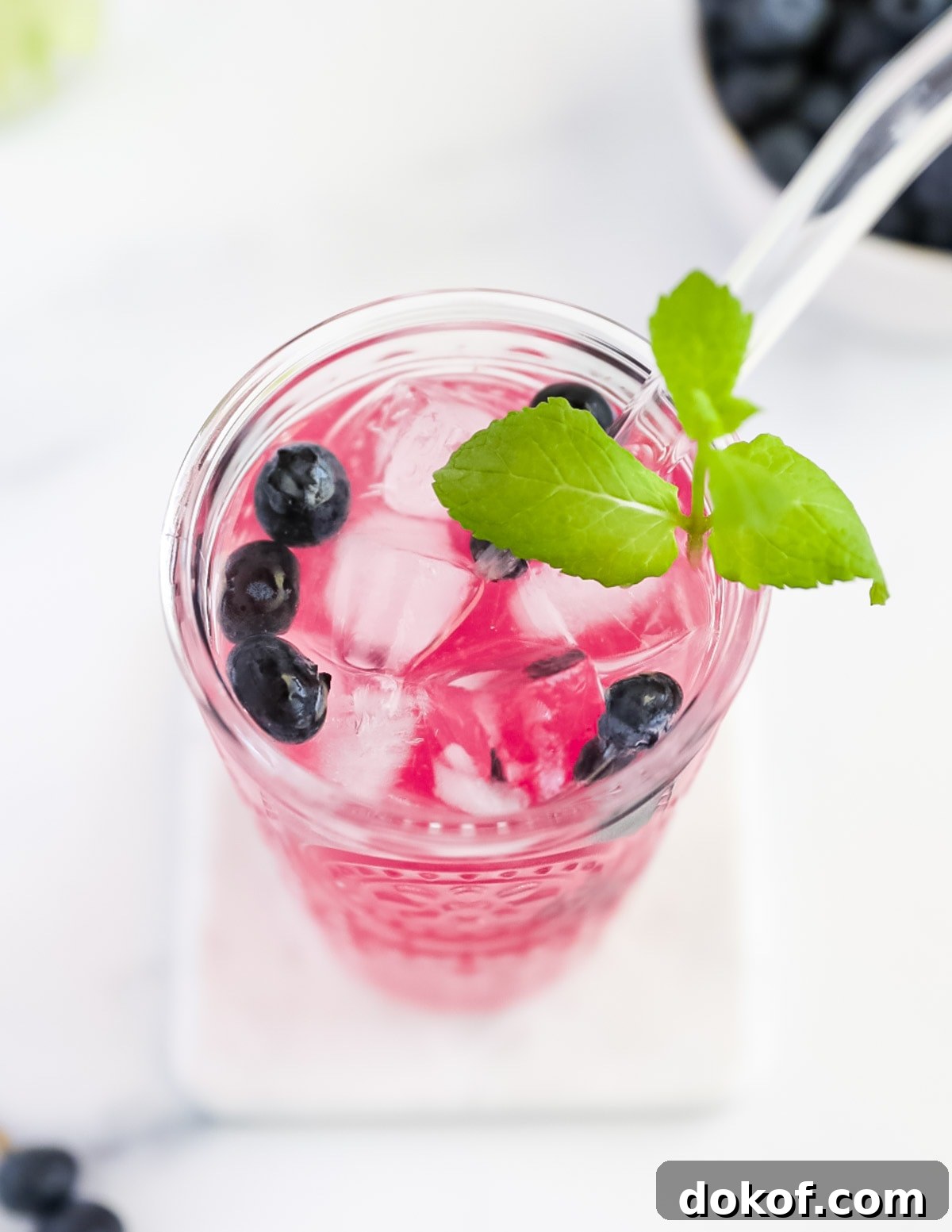 Refreshing Blueberry Lemonade Recipe with Fresh Lemon Zest 5 An overhead picture of a glass filled with bright pink lemonade. There are berries, mint, and ice cubes, along with a glass straw in the cup.