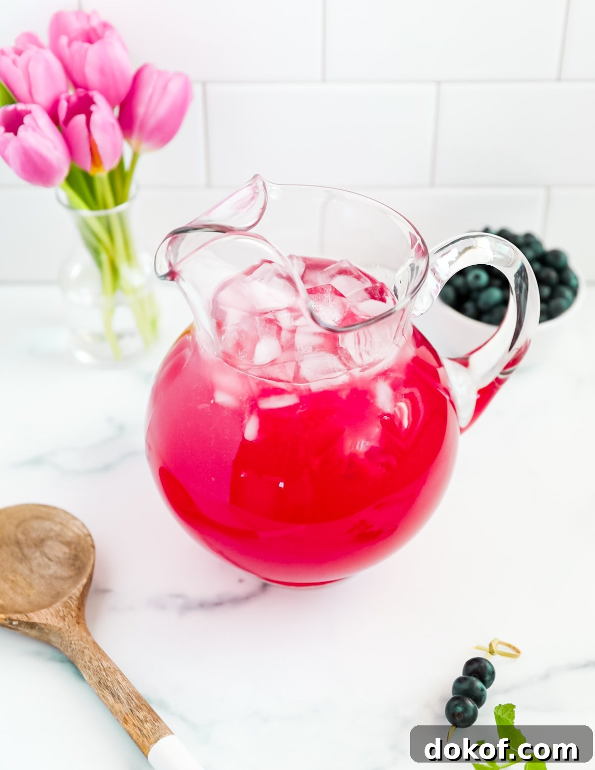 Refreshing Blueberry Lemonade Recipe with Fresh Lemon Zest 6 A clear glass pitcher filled with bright pink lemonade. There are ice cubes in the pitcher and flowers, berries, and a spoon outside