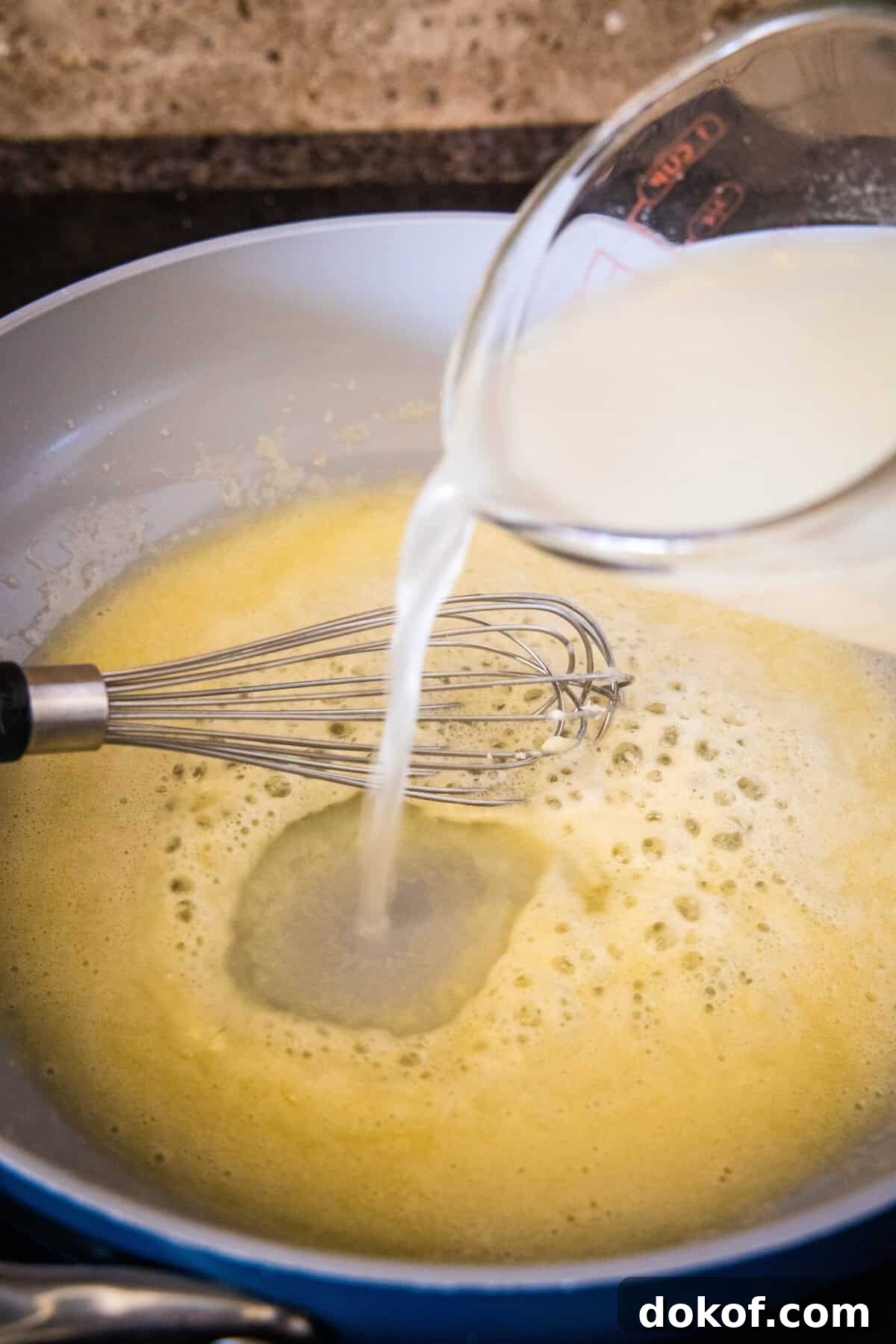Chicken broth being poured into a roux of butter and flour in a skillet.