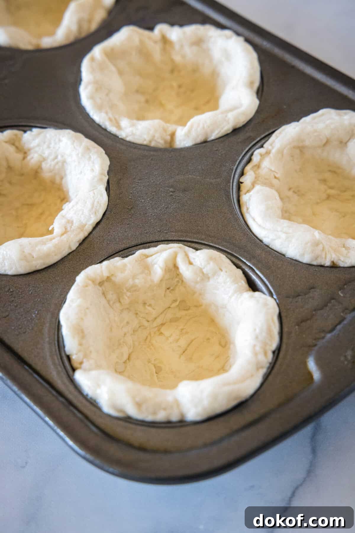Biscuit dough shaped into mini pie crusts inside the wells of a muffin tin.