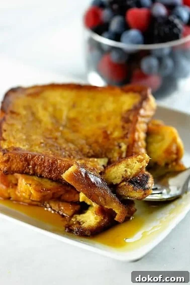 Photograph of French toast on a white plate with maple syrup and a bowl of fruit in the background.