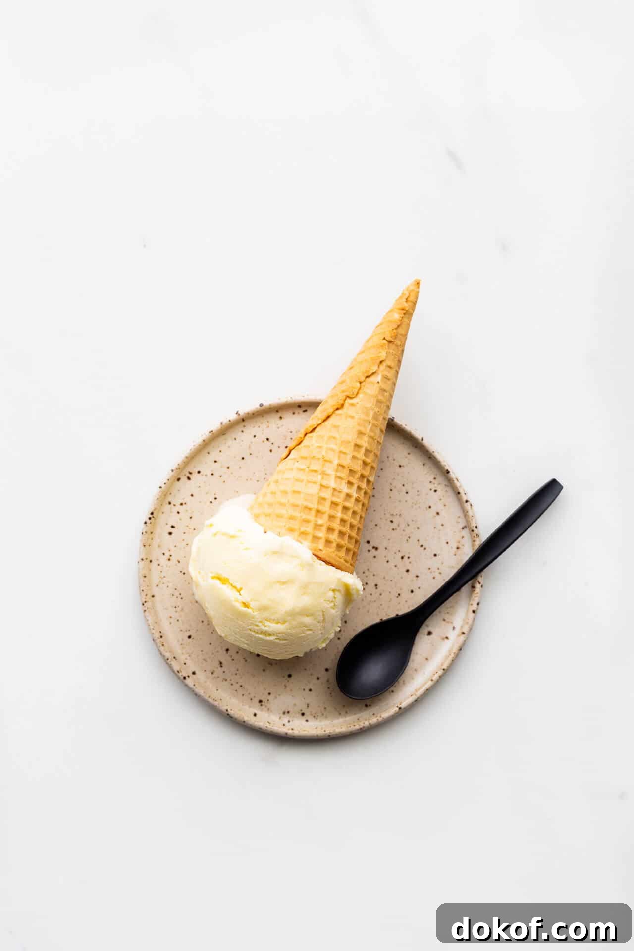 A scoop of lemon ice cream served on a speckled plate with a cone and a black spoon