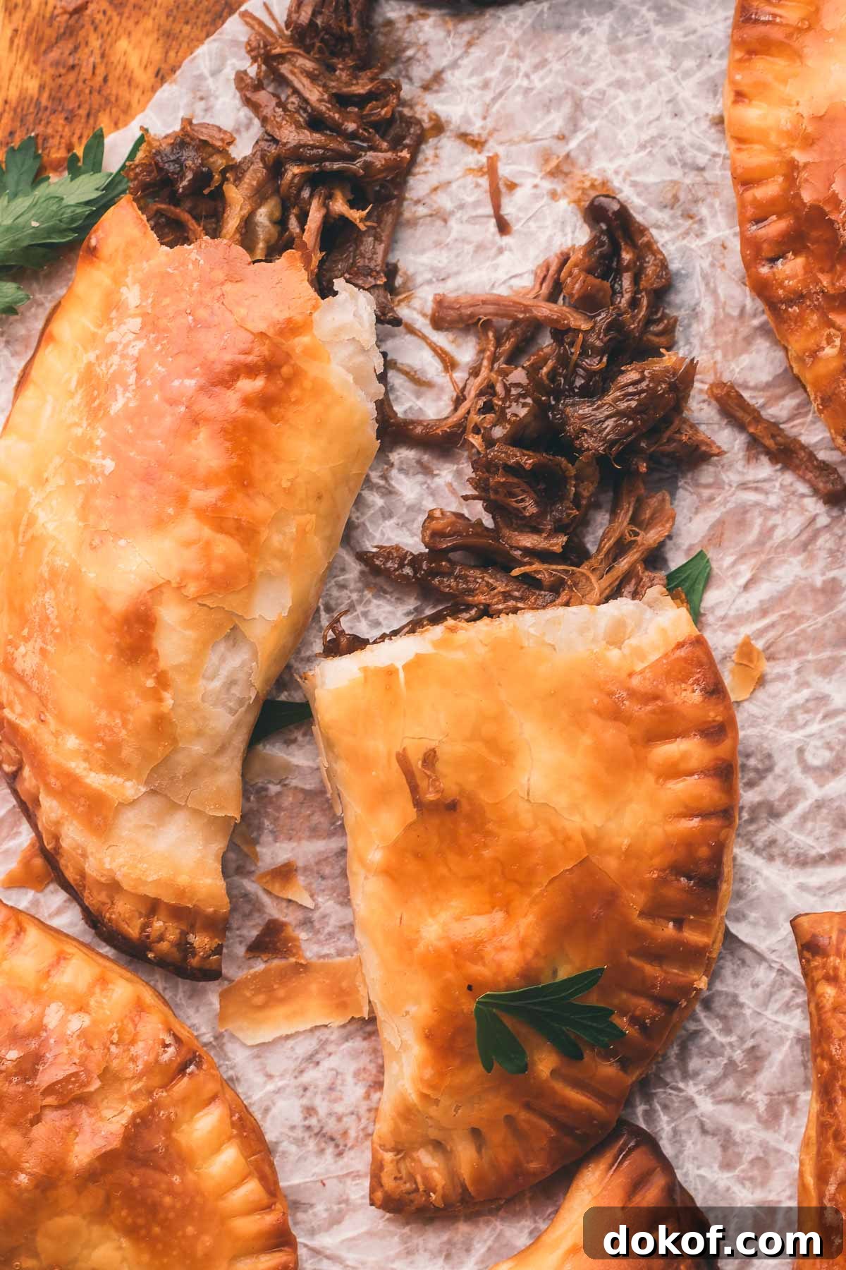 Golden brown stout beef hand pies are shown on parchment paper, one is broken open to reveal shredded beef filling. Parsley garnish is visible.
