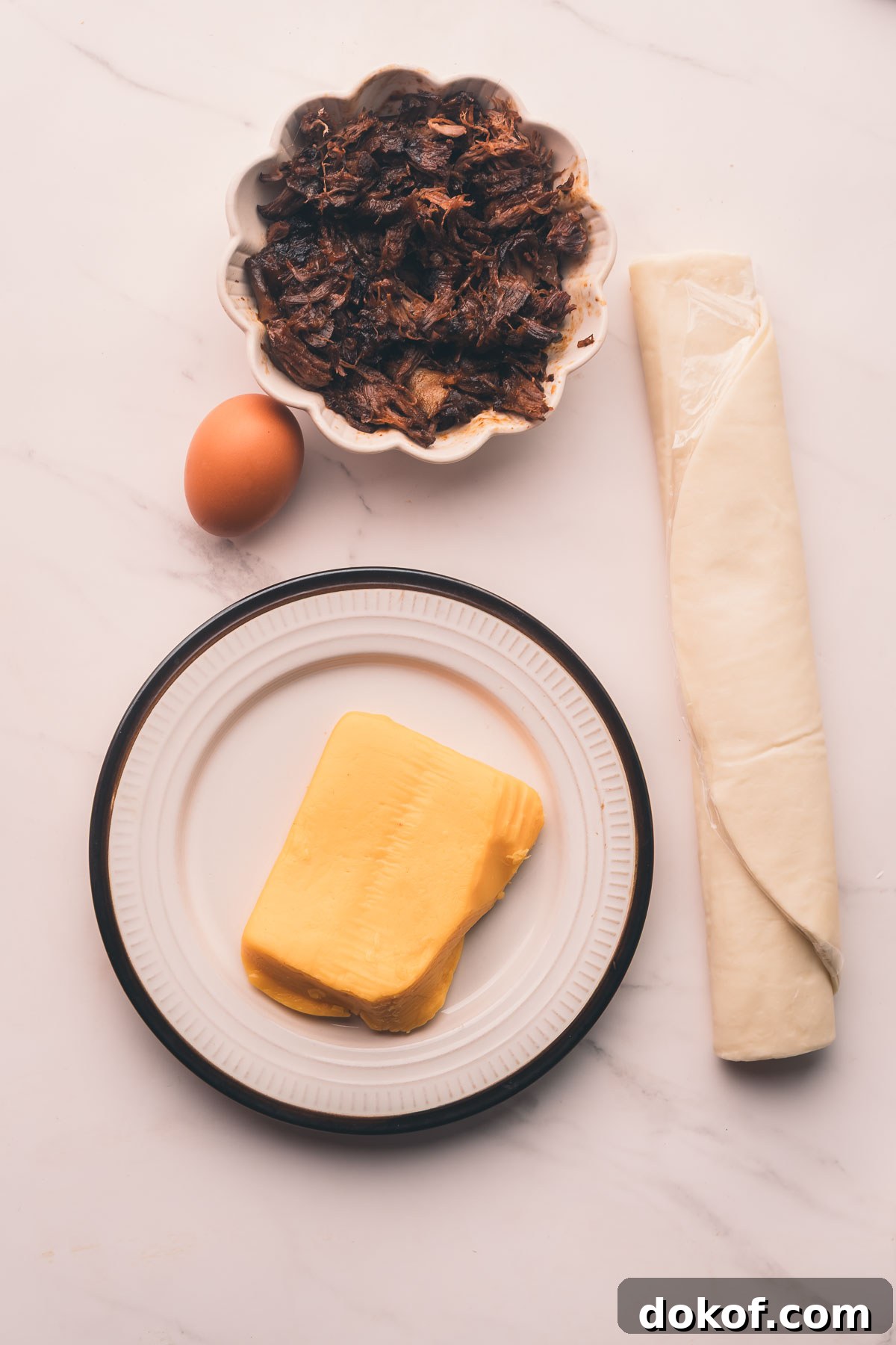 A plate with cheese, a raw egg, a bowl of shredded braised beef shanks, and a roll of puff pastry on a white marble surface.