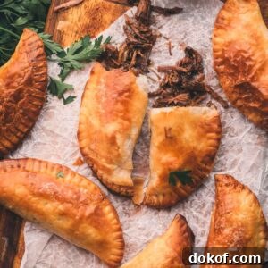 Five baked hand pies arranged on parchment paper, one split open to show shredded meat filling, with fresh parsley and a bowl of dark dipping sauce nearby.