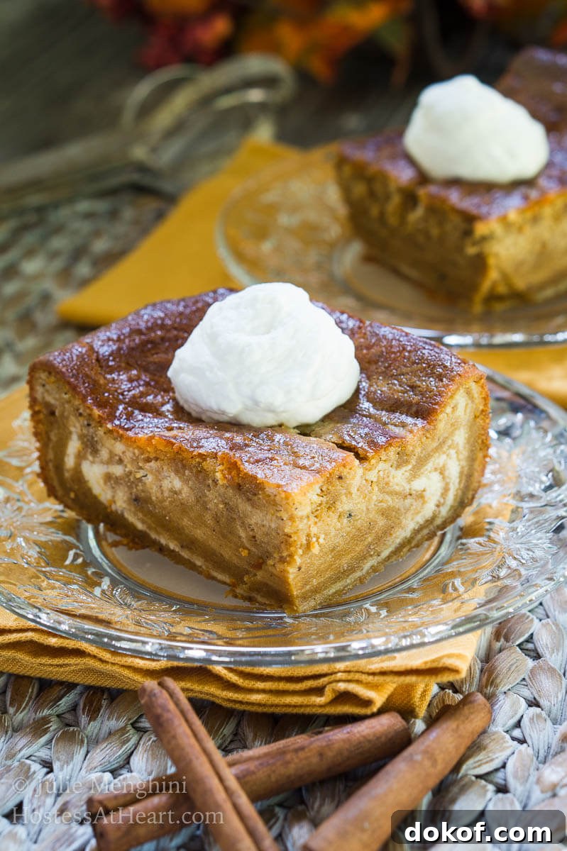 Creamy Pumpkin Cheesecake Swirl Bars Recipe 3 A piece of Pumpkin cheesecake with swirls of cream cheese filling running through it and topped with whipped cream sits on a glass plate over a gold napkin. A second slice sits in the background.
