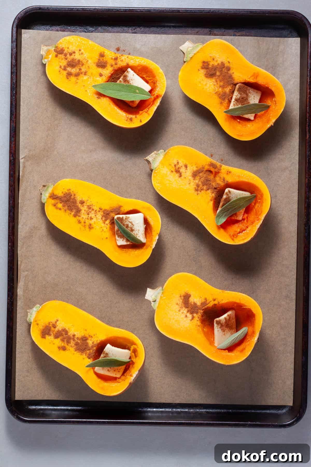 Halved honeynut squash on a parchment lined baking sheet with butter, cinnamon and sage in each cavity.