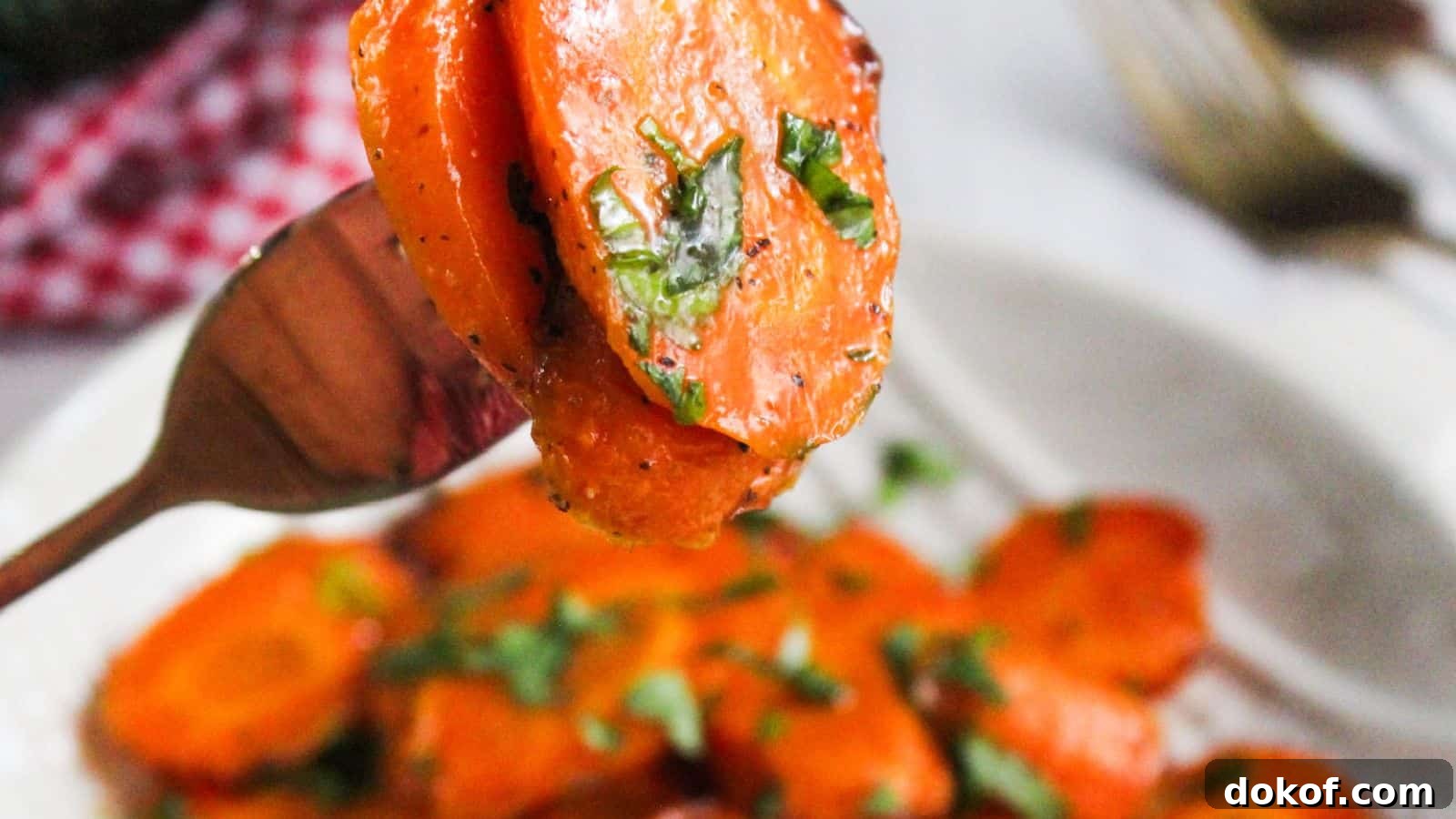 Sliced glazed carrots garnished with herbs on a fork, with more on a plate in the background.