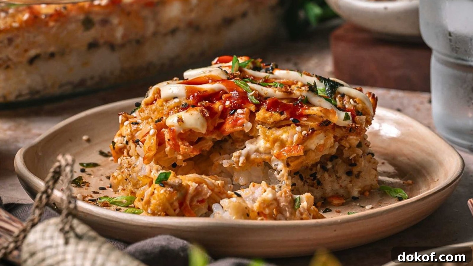 A serving of baked sushi casserole topped with sauces and herbs sits on a beige plate, with a portion removed to show layers of rice, crab, and creamy filling.